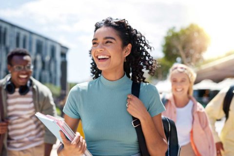 Three people walking outdoors near a building and trees, with one person carrying books and a backpack—suggesting student life or moving for studies.