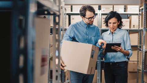 Two people in a warehouse surrounded by shelves and boxes, one holding a cardboard box and the other pointing at a tablet—suggesting organised storage or inventory management.