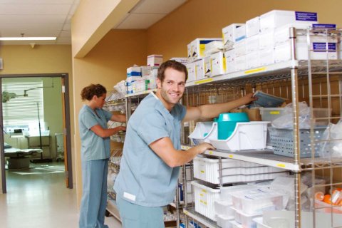 Two individuals in medical scrubs organizing supplies on metal shelves in a hospital storage area, with neatly arranged medical boxes and containers.
